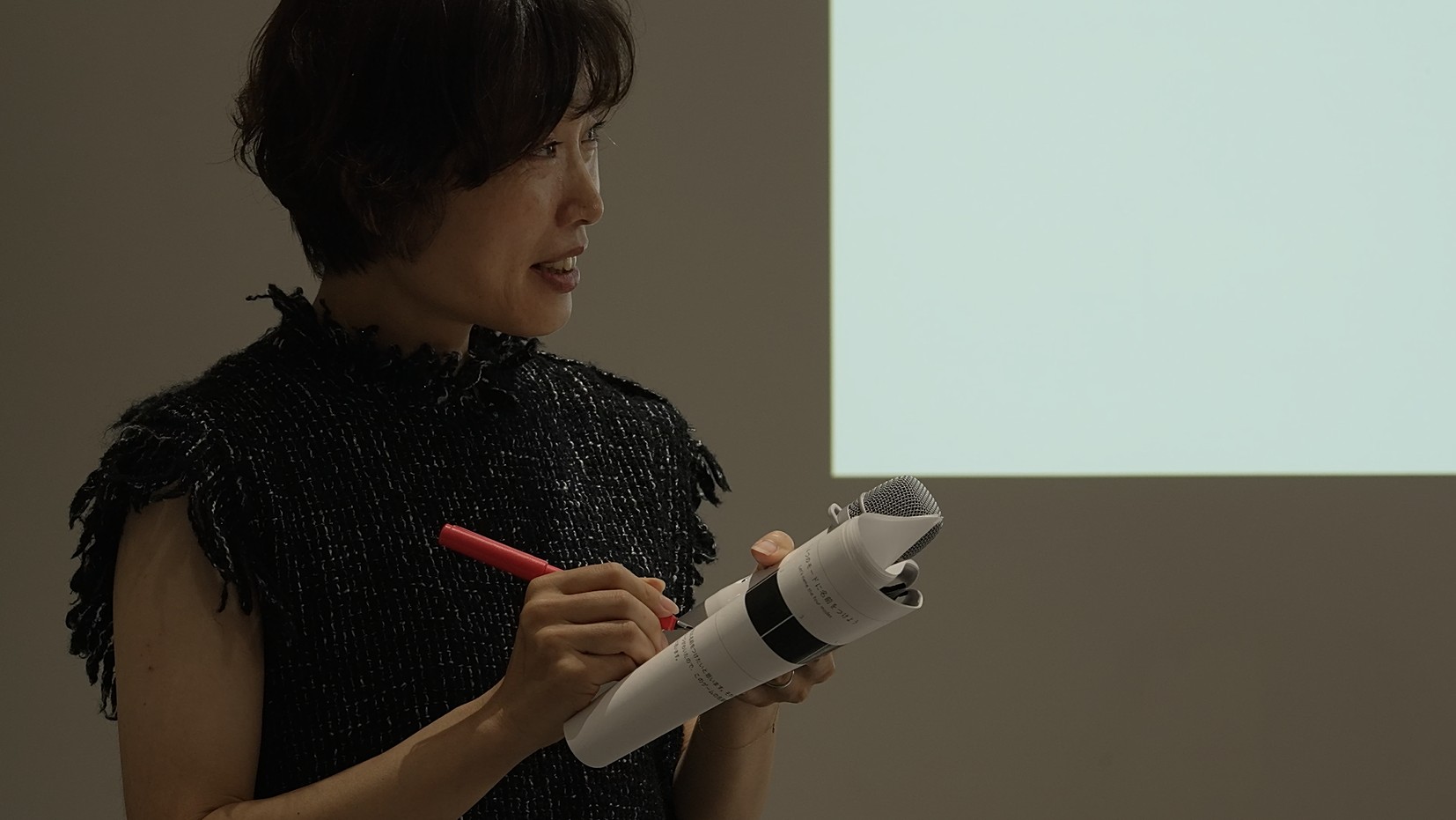 어두운 질감의 상의를 입은 짧은 머리의 여성이 프로젝터 화면 앞에 서서, 끝부분에 마이크 그릴이 결합된 원통형 종이 오브제를 한 손에 들고 다른 손에 든 빨간 펜으로 무언가를 적거나 표시하며 워크숍 참가자들에게 설명하고 있다.